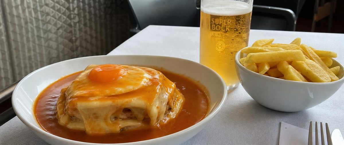 Francesinha in einer weißen Schale, übergossen mit Soße, dazu Pommes und ein Glas Super Bock